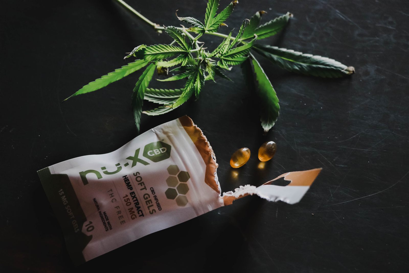 CBD softgel packaging, capsules, and hemp leaves arranged on a dark product photography surface.